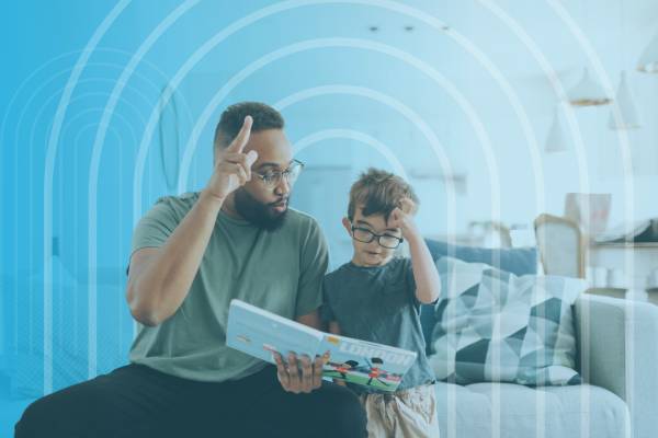 An adult and a young child sit together on a couch, looking at a tablet while using sign language to communicate.
